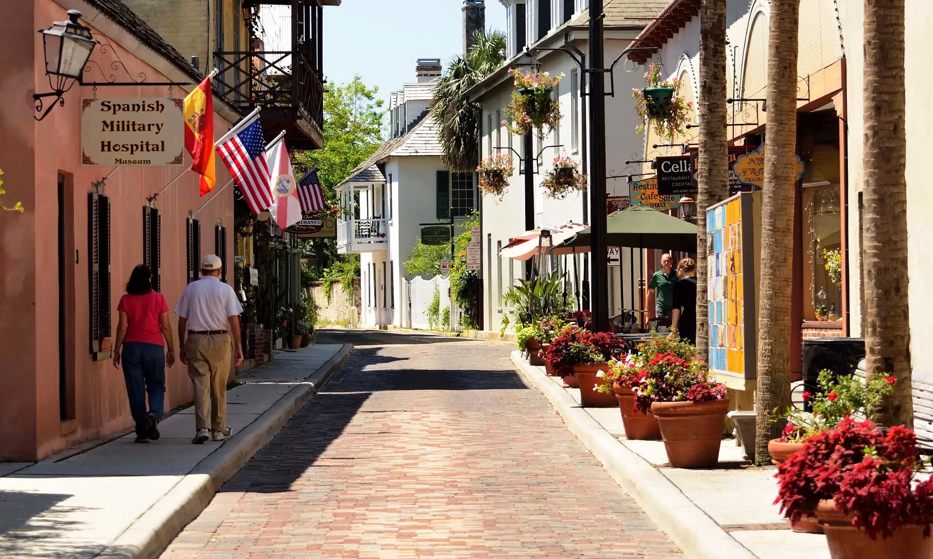 A sunlit cobblestone street lined with colorful flower pots and historic buildings, featuring flags and outdoor dining areas.